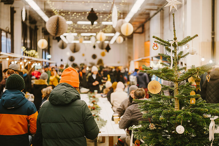 Blick in die winterlich geschmückte Alte Feuerwache am Flughafen Tempelhof. Im Vordergrund steht ein geschmückter Tannenbaum und im Hintergrund sieht man viele Menschen mit dicken Jacken und Mützen