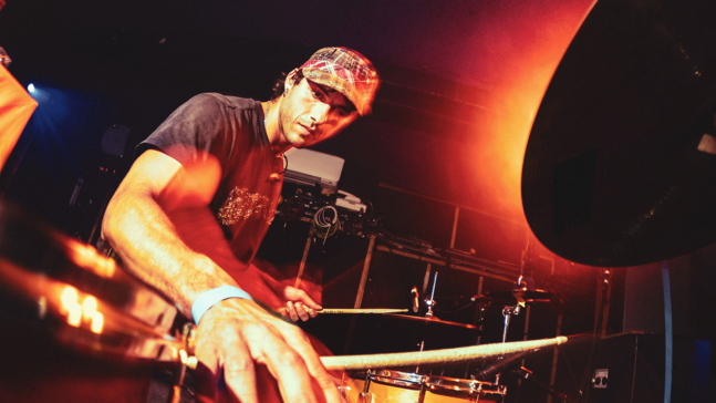 Oran Ray spielt auf einer Bühne ein Drumset. Die Szene ist dynamisch aus der Nähe aufgenommen; die Drumsticks sind in Bewegung leicht verwischt. Warmes, orangefarbenes Bühnenlicht fällt von der Seite auf den Musiker und das Schlagzeug, im Hintergrund sind Kabel und Bühnentechnik erkennbar.