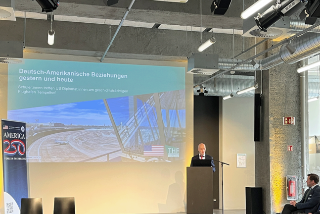 Alan Meltzer im Besucherzentrum am Flughafen Tempelhof vor einer Leinwand  auf der "Deutsch-Amerikanische Beziehungen gestern und heute" steht