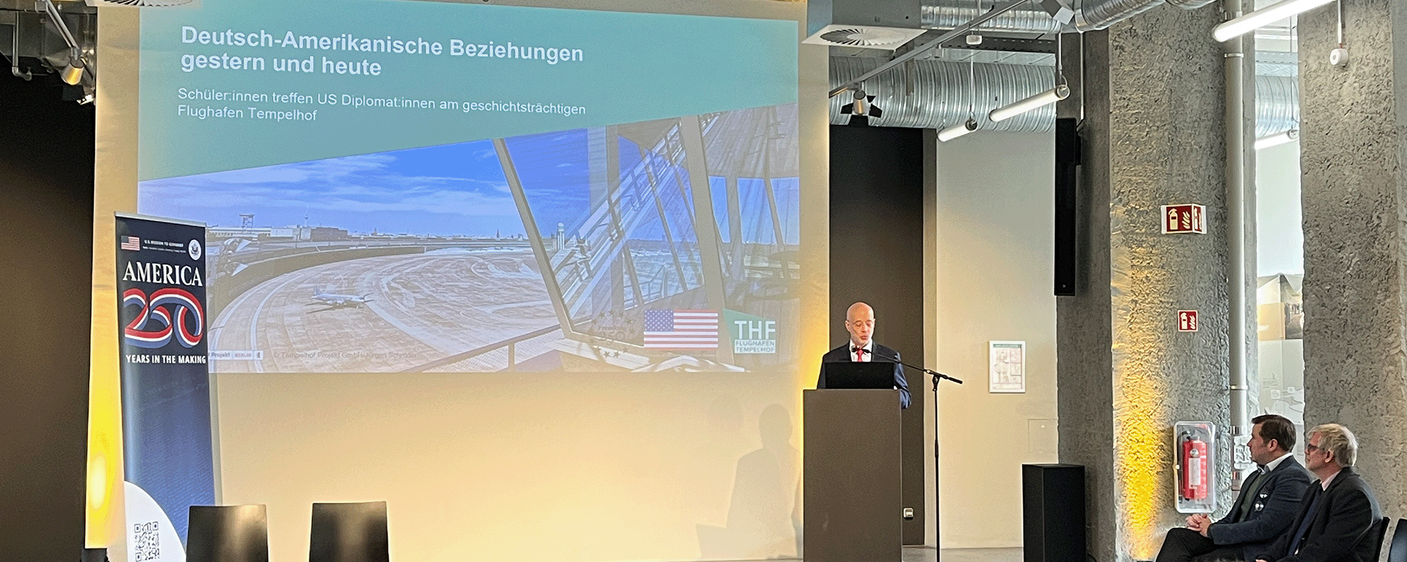 Alan Meltzer im Besucherzentrum am Flughafen Tempelhof vor einer Leinwand  auf der "Deutsch-Amerikanische Beziehungen gestern und heute" steht