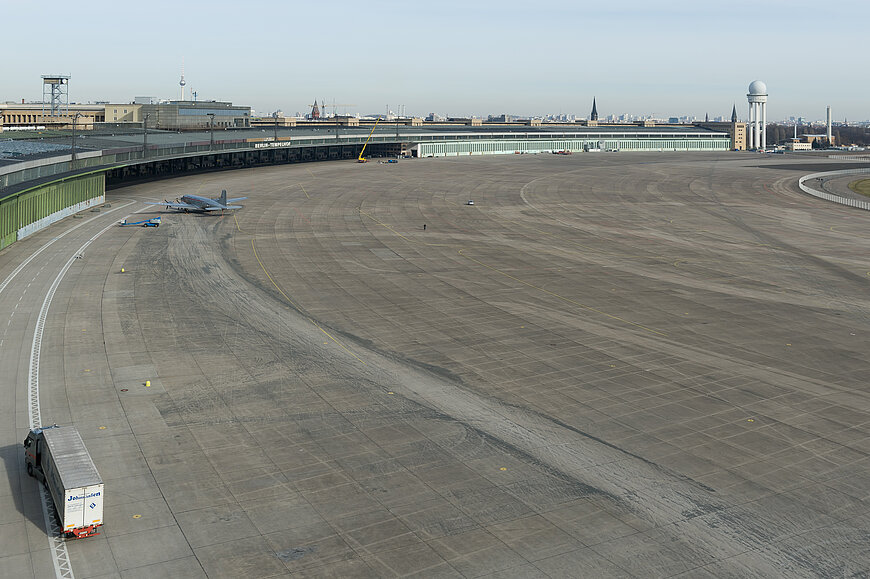 Gesamtansicht desVorfeldes des Flughafen Tempelhof. Links fährt ein weißes Fahrzeug. Im Hintergrund erstreckt sich das lange, dunkle Hauptgebäude des Flughafens mit einem Kontrollturm am rechten Ende.