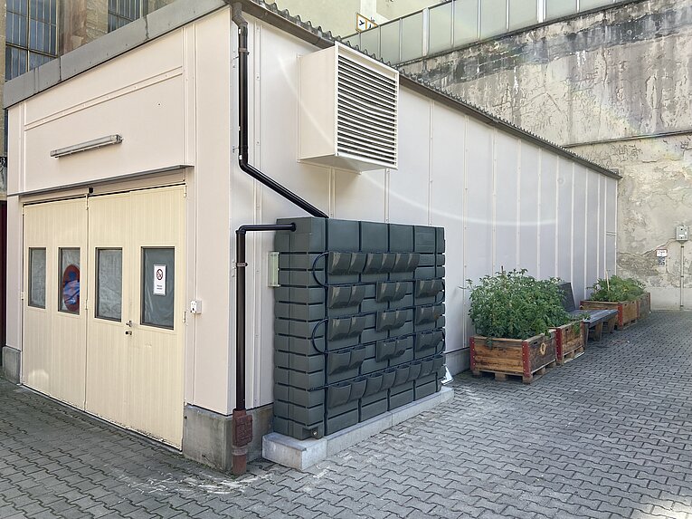 Hochbeete und Wassertank bei der Fliegerwerkstatt vor einer weißen Wand