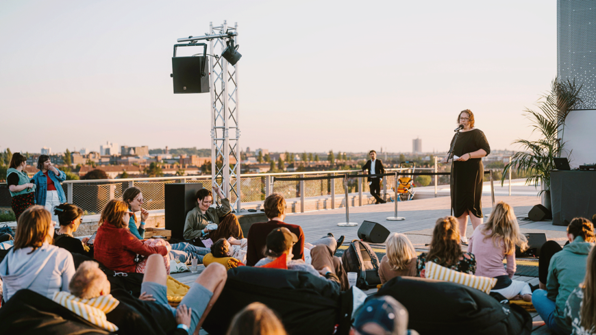 Eine Person steht auf einer Bühne auf der Dachterrasse des THF TOWER. Einige Menschen sitzen davor in Sandsäcken und hören zu.