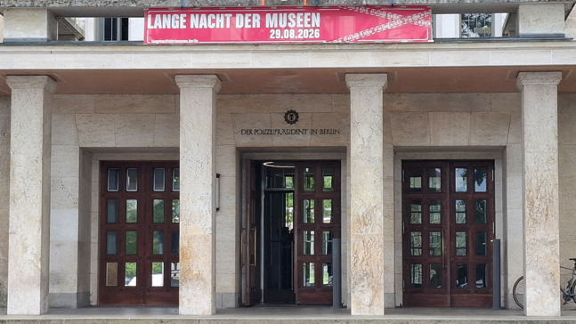Außenfassade des Polizeigebäudes mit drei großen Holztüren und Säulen am Eingang; darüber ein rotes Banner zur Langen Nacht der Museen 29.08.2026. Über der mittleren Tür steht die Inschrift Der Polizeipräsident in Berlin. Rechts neben dem Eingang steht ein Fahrrad.