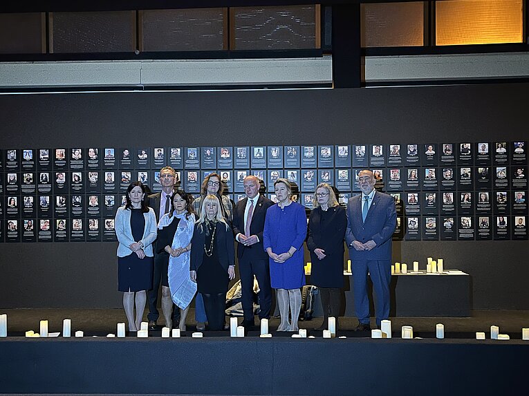 Der Senat von Berlin steht vor der Gedenkwand in der Nova-Ausstellung am Flughafen Tempelhof. Im Hintergrund hängen Fotos von den Geiseln, im Vordergrund leuchten Kerzen