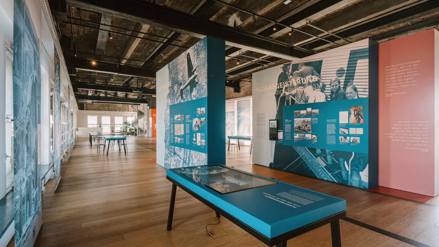 Ausstellungsraum im THF TOWER mit Holzfußboden, großen Informations- und Grafiktafeln zu Luftfahrtgeschichte (u. a. „Flugbegeisterung“ und „Lufthansa“) sowie Tischen und interaktiven Stationen; links fällt Tageslicht durch hohe Fenster in den Raum.