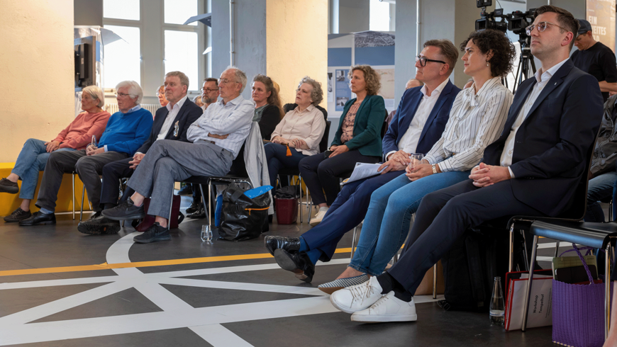 Ein Publikum sitzt im Besucherzentrum am Flughafen Tempelhof auf Stühlen und schaut in eine Richtung