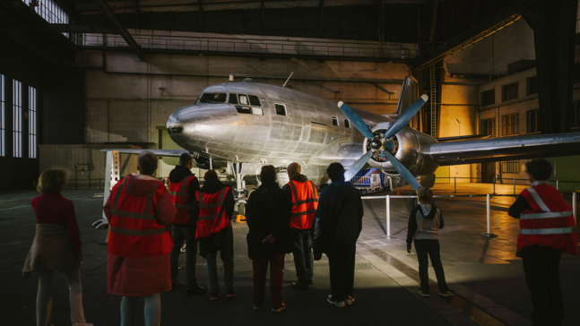 Gruppe von Besucherinnen und Besuchern in roten Sicherheitswesten steht bei der Familiennacht in einem großen Hangar am Flughafen Tempelhof vor einem Flugzeug mit. Die Halle ist hoch, industriell beleuchtet und hat große Fenster entlang der Seiten.