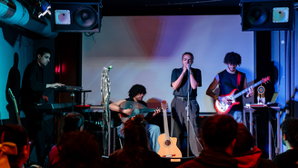 Kleine Bühne mit vier Musikerinnen und Musikern, beleuchtet in Blau‑ und Lilatönen; eine Sängerin steht am Mikrofon, begleitet von Keyboard, Akustik‑ und E‑Gitarre, während das Publikum im Vordergrund sitzt und zuhört.