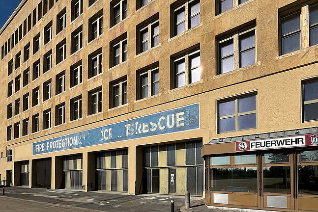 Die Fassade der Alten Feuerwache mit ihrem hellen, leicht gealterten Steinbau und den regelmäßig angeordneten Fenstern. Im unteren Bereich sind große Toröffnungen und verblasste Beschriftungen wie „Fire Protection … Rescue“ sowie ein klar lesbares „Feuerwehr“-Schild zu sehen.