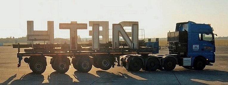 Die Leuchtbuchstaben werden mit einem LKW abtransportiert. Im Hintergrund geht die leuchtende Sonne auf