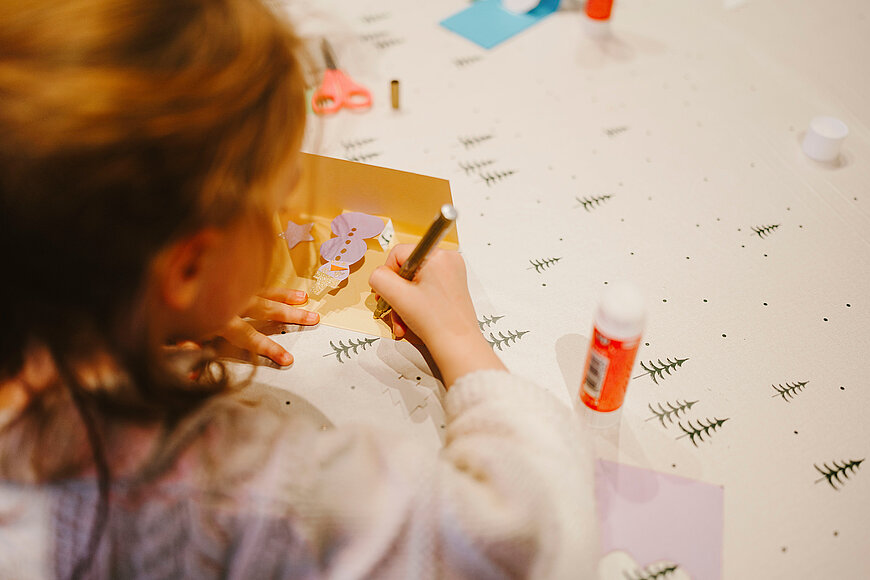 Ein junges Mädchen von hinten, dass eine Pop-up-Karte ausmalt. Neben ihr steht ein Kleber auf dem tisch