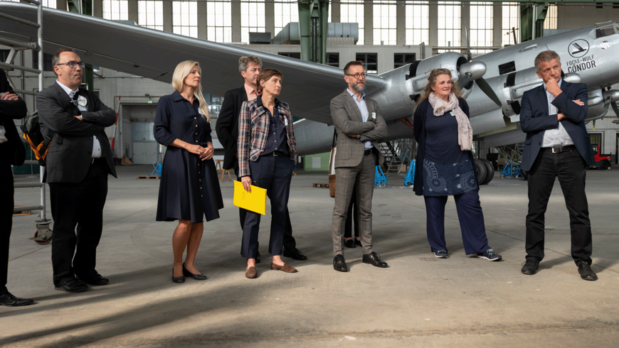 Eine Gruppe von Menschen in Bussines-Outfits steht vor einem historischen Flugzeug im Hangar am Flughafen Tempelhof