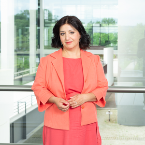 Porträt von Yalda Zarbakhch in einem korall-rotem Kleid und Jacke vor einer Glaswand