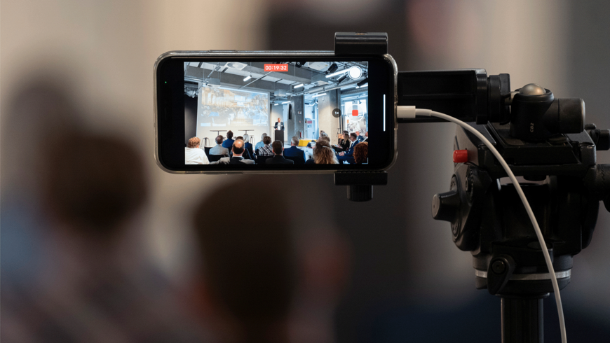 Foto von einem Handy in einem Stativ, dass eine Konferenz aufzeichnet
