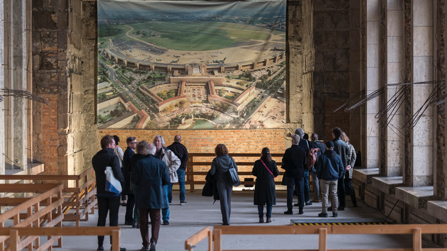 Eine Gruppen von Menschen in einer große, historische Eingangshalle mit hohen Säulen vor einer Luftaufnahme des Flughafen Tempelhof 