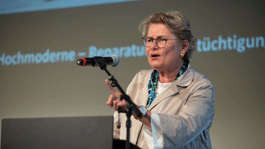 Prof. Petra Kahlfeldt auf einer Bühne mit Mikrophon. Im Hintergrund eine Leinwand