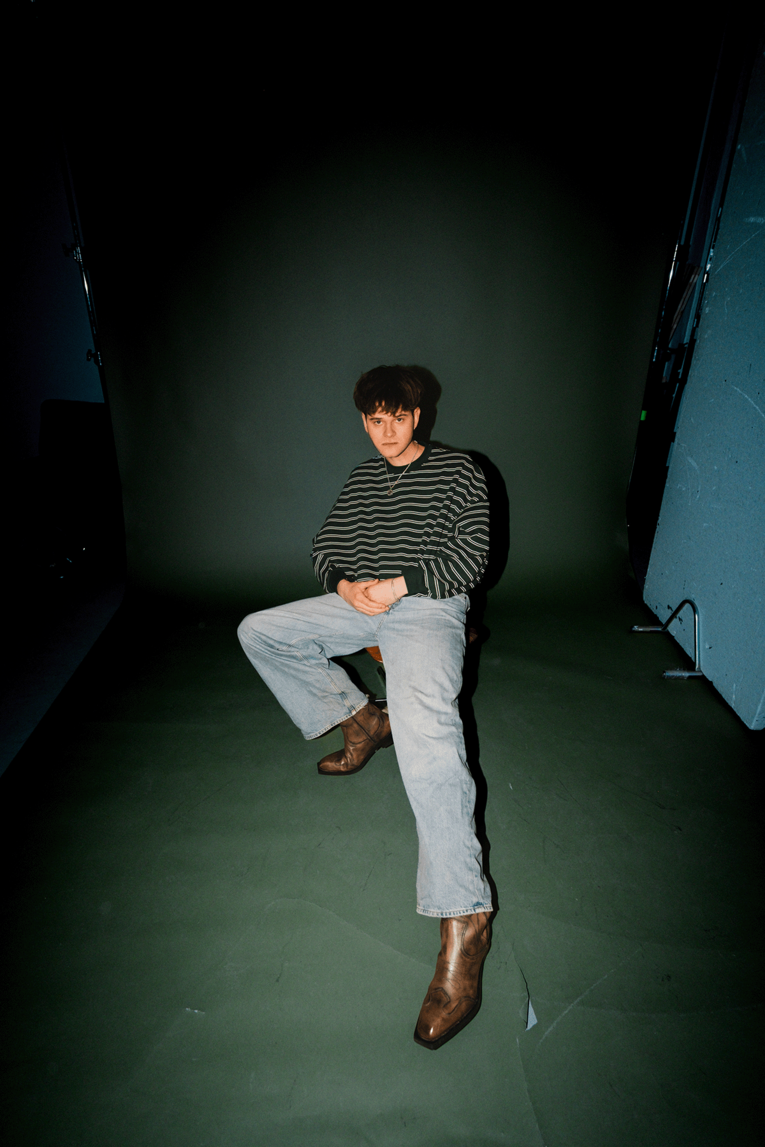 Porträt des Musikers JAS in einem Studio vor dunkelgrünem Hintergrund. Er sitzt auf einem Hocker, trägt ein schwarz‑weiß gestreiftes Langarmshirt, hellblaue Jeans und braune Stiefel. Mit kurz geschnittenem dunklem Haar blickt er ruhig in die Kamera, die Hände locker vor dem Körper verschränkt. Das Licht hebt ihn klar vom gedimmten Hintergrund ab.