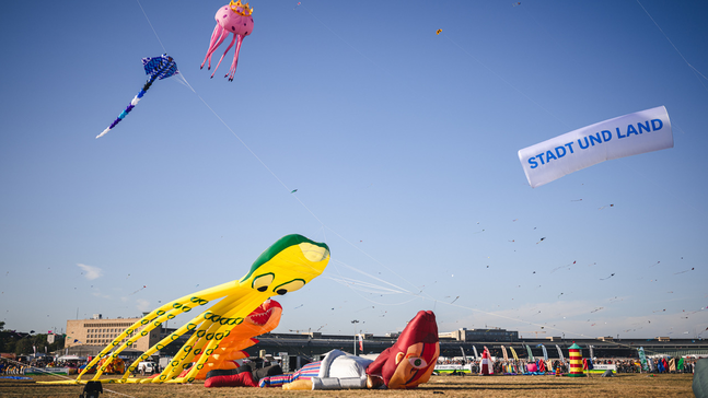Bunte Riesendrachen mit blauem Himmel auf dem Tempelhofer Feld. Im Hintergrund ist das Flughafengebäude zu sehen