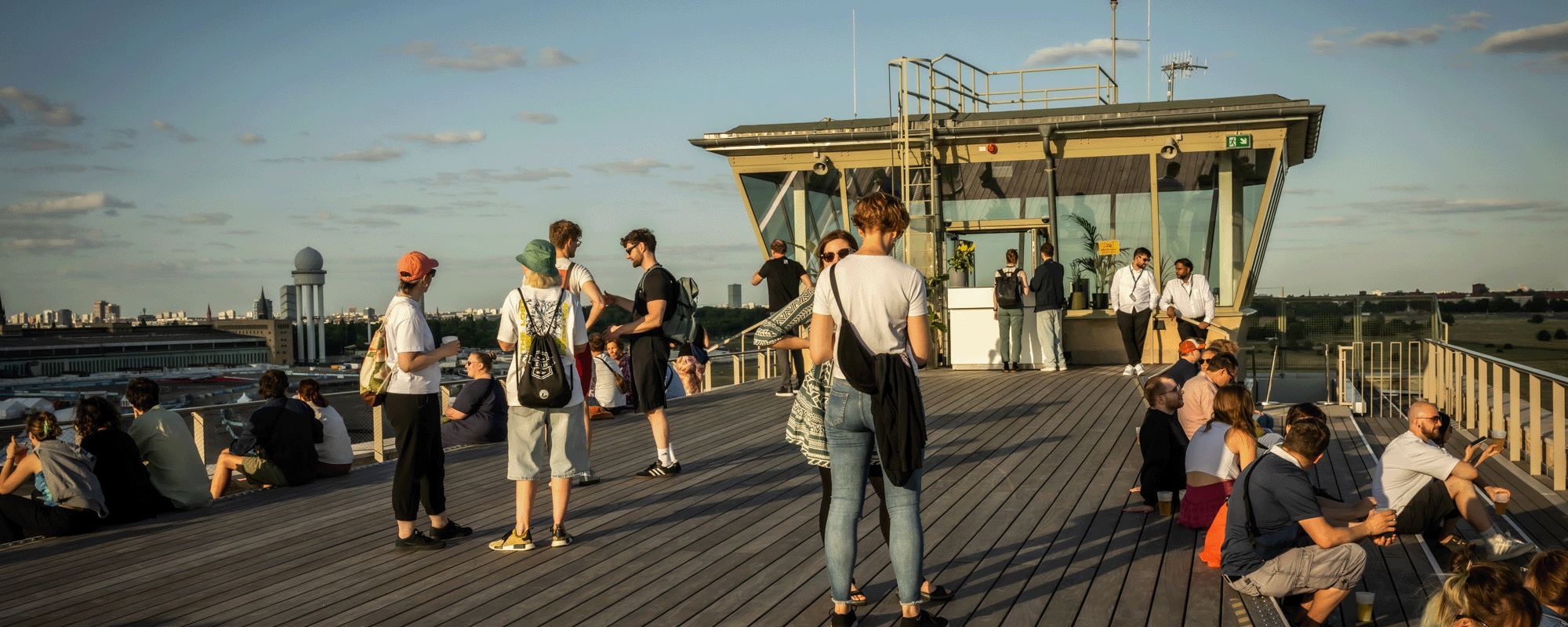 Eine größere Gruppe von Menschen steht und sitzt auf der weitläufigen Holzterrasse des THF TOWER in warmem Sonnenlicht. Im Hintergrund ragt der Tower empor, daneben ist eine städtische Skyline mit markanten Gebäuden und einem Radom zu sehen. Die Personen wirken entspannt, unterhalten sich oder blicken über die Stadt.