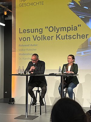 Man sieht Volker Kutscher und die Moderatorin auf der Bühne sitzen. 