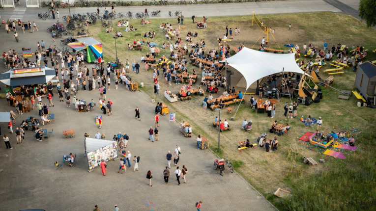Eine weit gefasste Vogelperspektive auf den Neulich Biergarten am THF TOWER. Das Areal teilt sich in einen gepflasterten Bereich mit Foodtrucks, bunten Verkaufsständen und Warteschlangen sowie eine angrenzende Grünfläche.