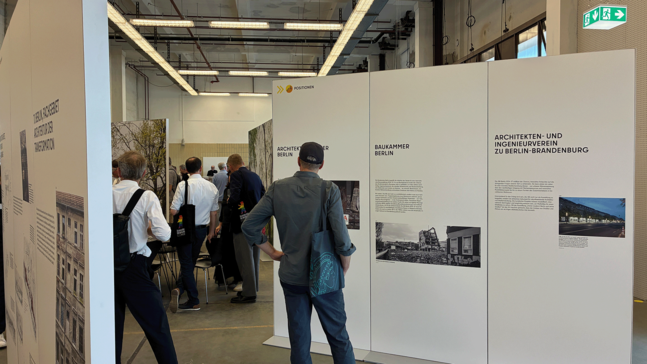 Eine moderne Ausstellungshalle zur IBA in der Alten Feuerwache mit mehreren Besucherinnen und Besuchern, die vor großen weißen Infotafeln stehen. Die Tafeln zeigen Texte und historische sowie aktuelle Fotos zu Architekturorganisationen in Berlin und Brandenburg.