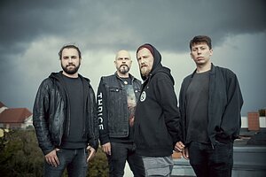 Vier Männer, in dunkler Kleidung, stehen auf einer Dachterrasse. Der Himmel über Ihnen ist grau und wolkig.