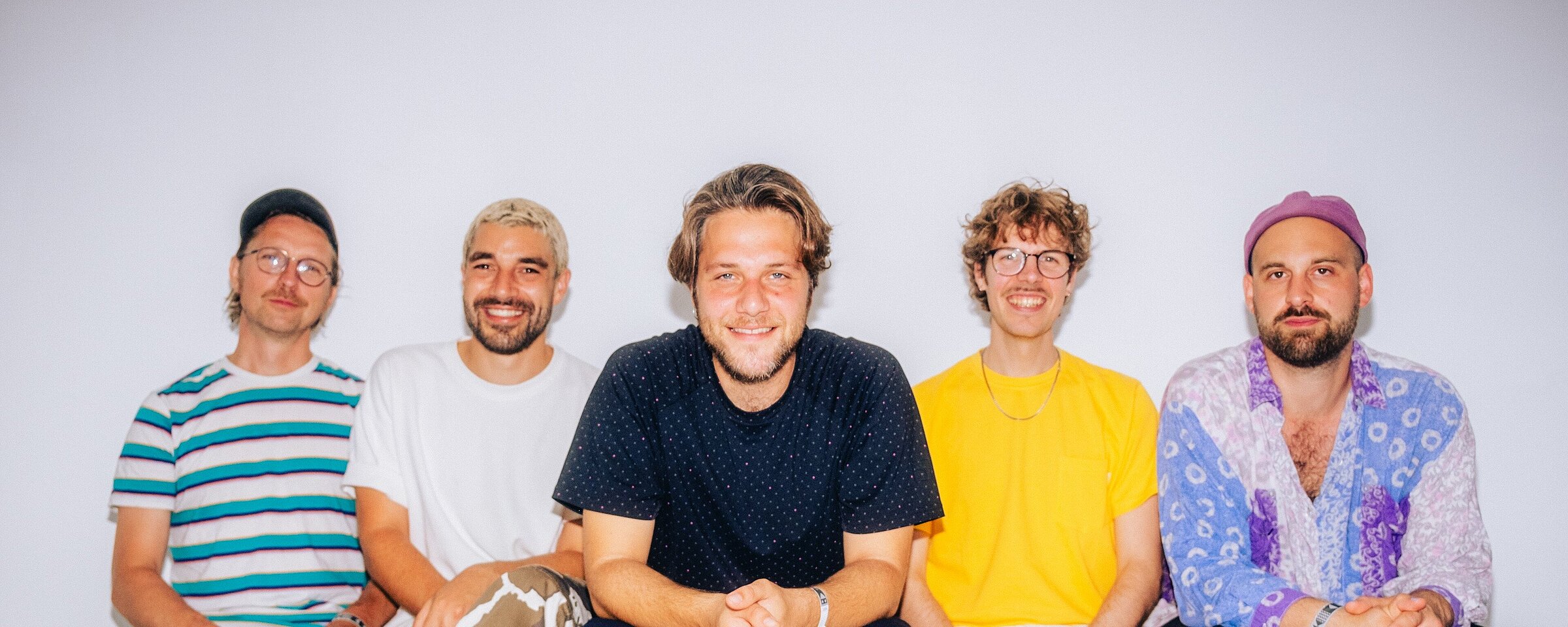 Zu sehen sind fünf junge Bandmitglieder der Band IL  CIVETTO, die in einer Rehe sitzen. Sie tragen bunte Klamotten ud lächeln. 