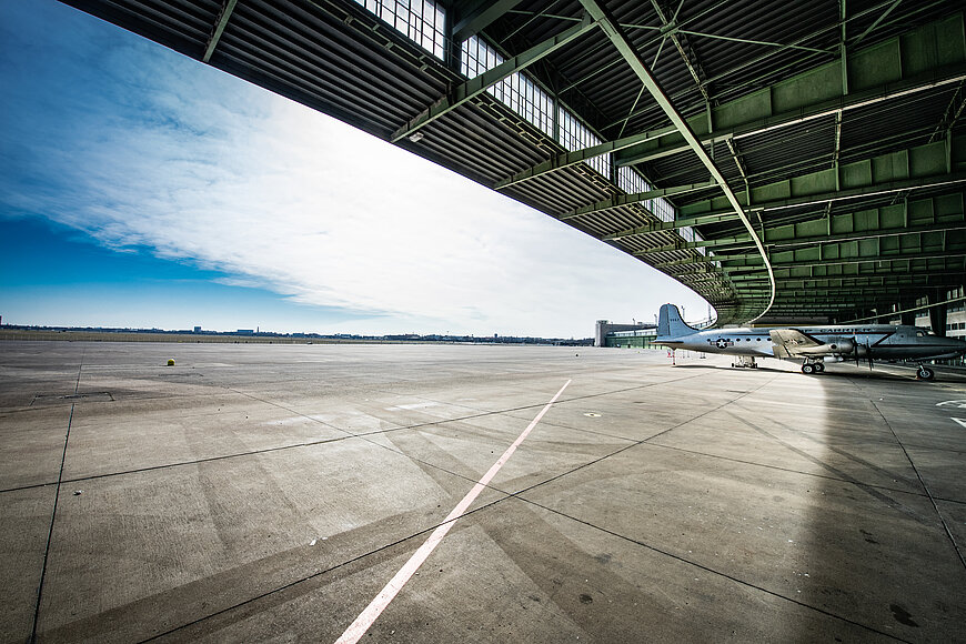 Blick unter die bogenförmige Stahlkonstruktion eines Hangardachs hinaus auf das weite, sonnige Rollfeld. Im HIintergrund ein Flugzeug