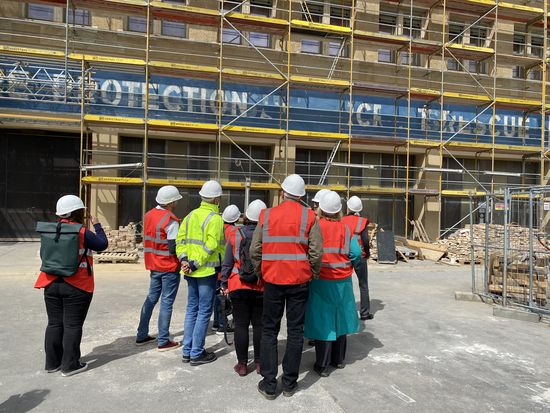 Besuchergruppe mit roten Warnwesten und weißen Bauhelmen steht vor einem Gebäude mit Baugerüst