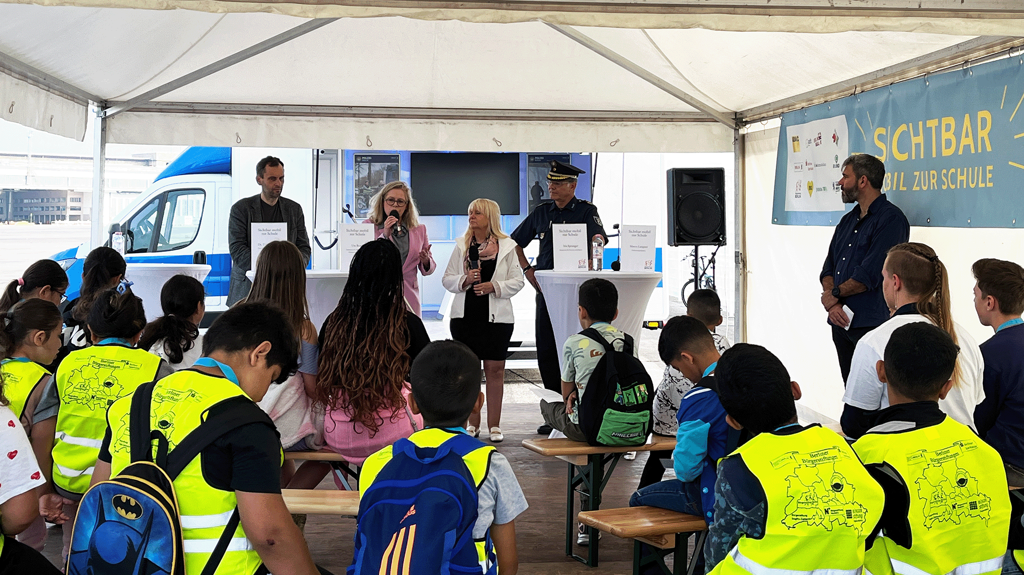 Kinder mit gelben Warnwesten sitzen bei einer Schüler:innenpressekonferenz beim Aktionstag Sichtbar Mobil zur Schule. vorne stehen Ute Bonde, Senatorin für Mobilität, Verkehr, Klimaschutz und Umwelt, Iris Spranger, Senatorin für Inneres und Sport, Dr. Torsten Kühne, Staatssekretär für Schulbau und Schuldigitalisierung, Dr. Andrea Grebe, Vorstandsvorsitzende der Bürgerstiftung Berlin und Polizeivizepräsident Marco Langner. 