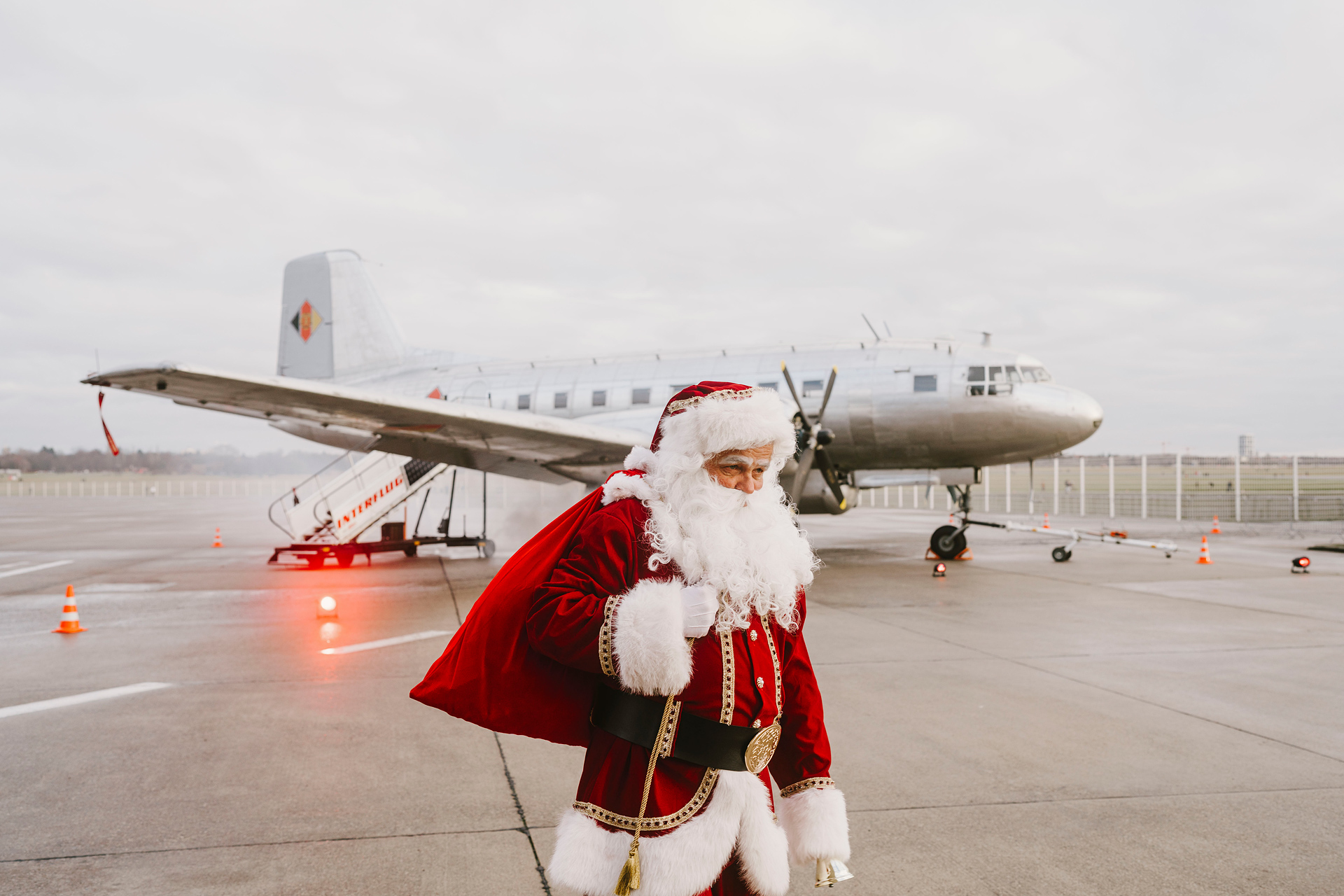 Ein verkleideter Weihnachtsmann mit einem großen roten Sack auf dem Rücken läuft am Flughafen Tempelhof vor einem Rosinenbomber-Flugzeug