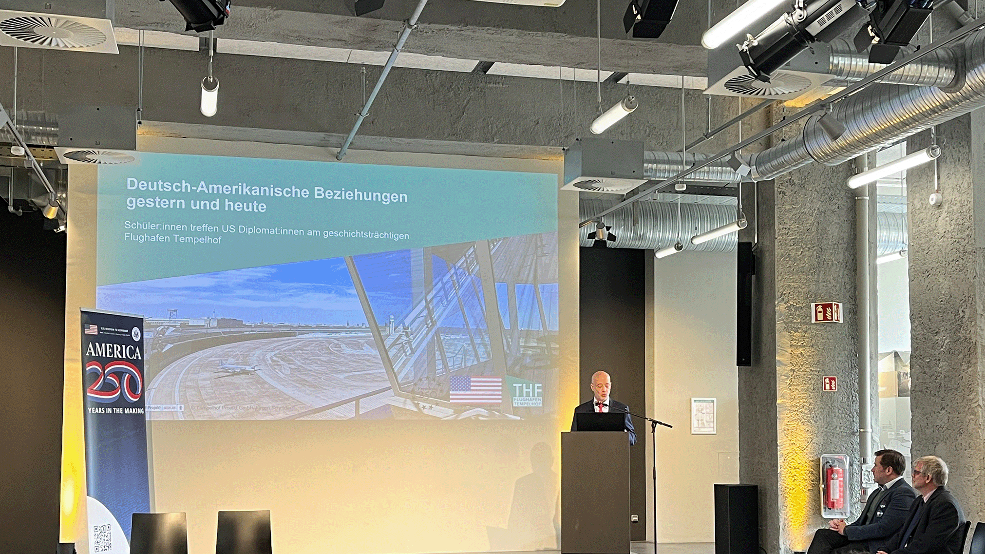 Alan Meltzer im Besucherzentrum am Flughafen Tempelhof vor einer Leinwand  auf der "Deutsch-Amerikanische Beziehungen gestern und heute" steht
