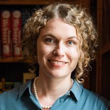 Porträt von Carolin Liebisch-Gümüş mit lockigem, hellblondem Haar, die ein türkisfarbenes Oberteil und eine Perlenkette trägt. Im Hintergrund stehen Bücherregale voller Bücher, einige mit roten Einbänden und goldener Schrift, was eine studienartige, ruhige Atmosphäre erzeugt