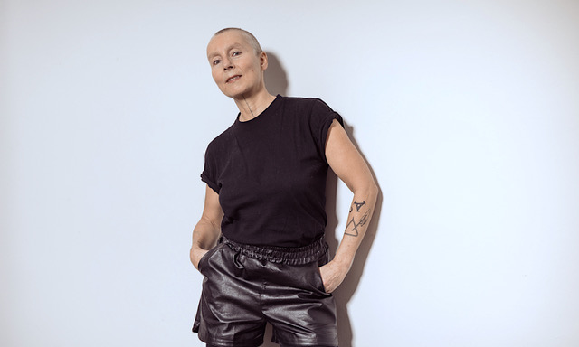 DJ Femalemacho mit rasiertem Kopf steht vor einer hellen, glatten Wand. Sie trägt ein schwarzes T‑Shirt und schwarze Leder-Shorts, die Hände locker in den Taschen. Auf dem linken Unterarm ist ein geometrisches Tattoo sichtbar. Die Szene ist weich ausgeleuchtet und wirkt minimalistisch.