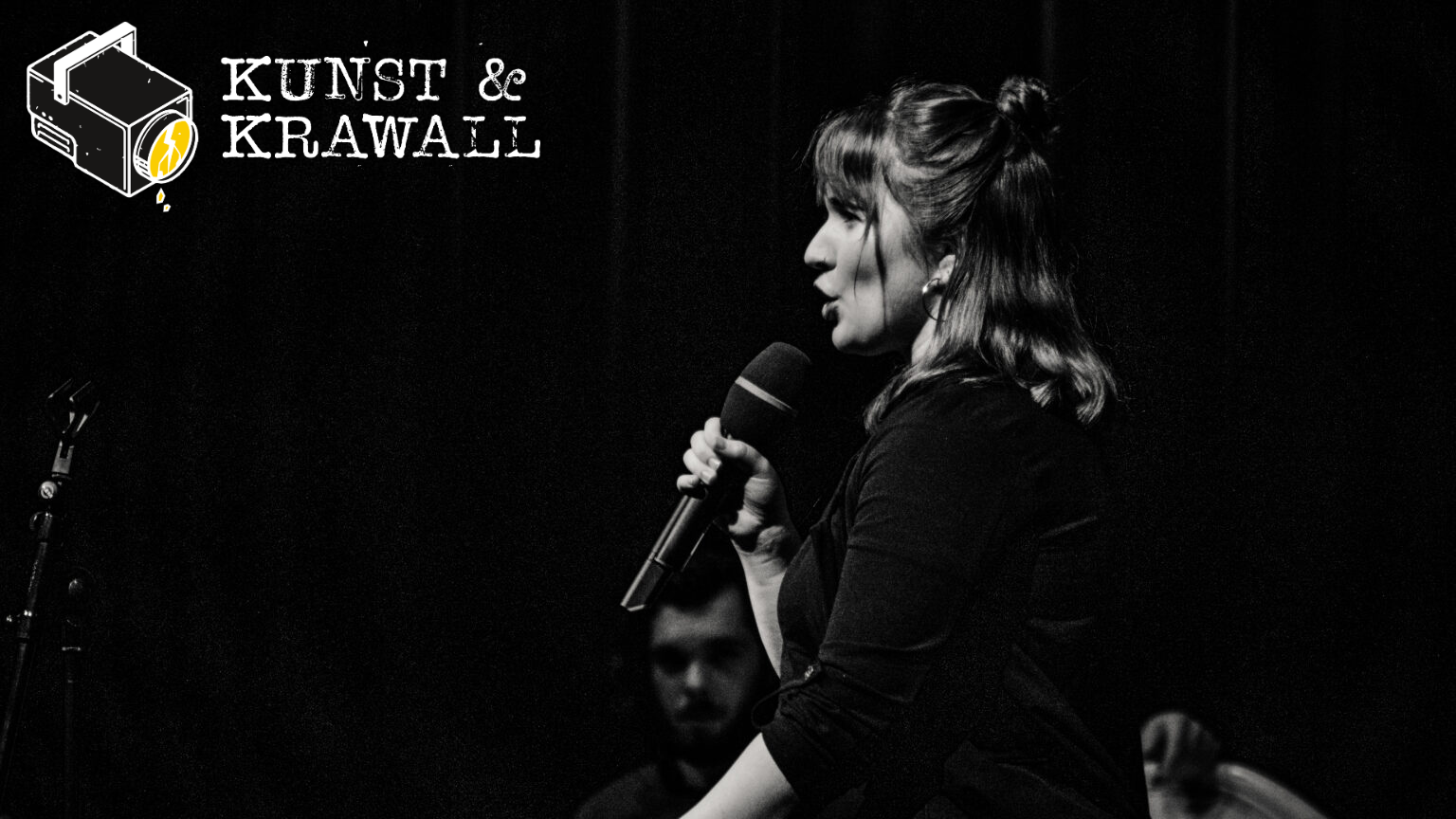 Ein dunkles Foto von einer Frau, die auf einer Bühne steht und in ihrer Hand ein Mikrophon hält. Oben links steht "Kunst &  Krawall" und ein gezeichneter Scheinwerfer