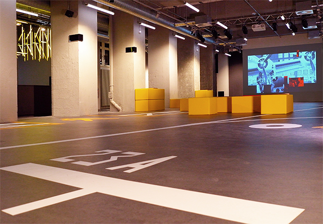 Blick in den Innenraum des Besucherzentrums CHECK IN am Flughafen Tempelhof. Ein Beamer strahlt ein Bild eines Flugzeugs an die Wand, gelbe Sitzkissen stehen im Raum