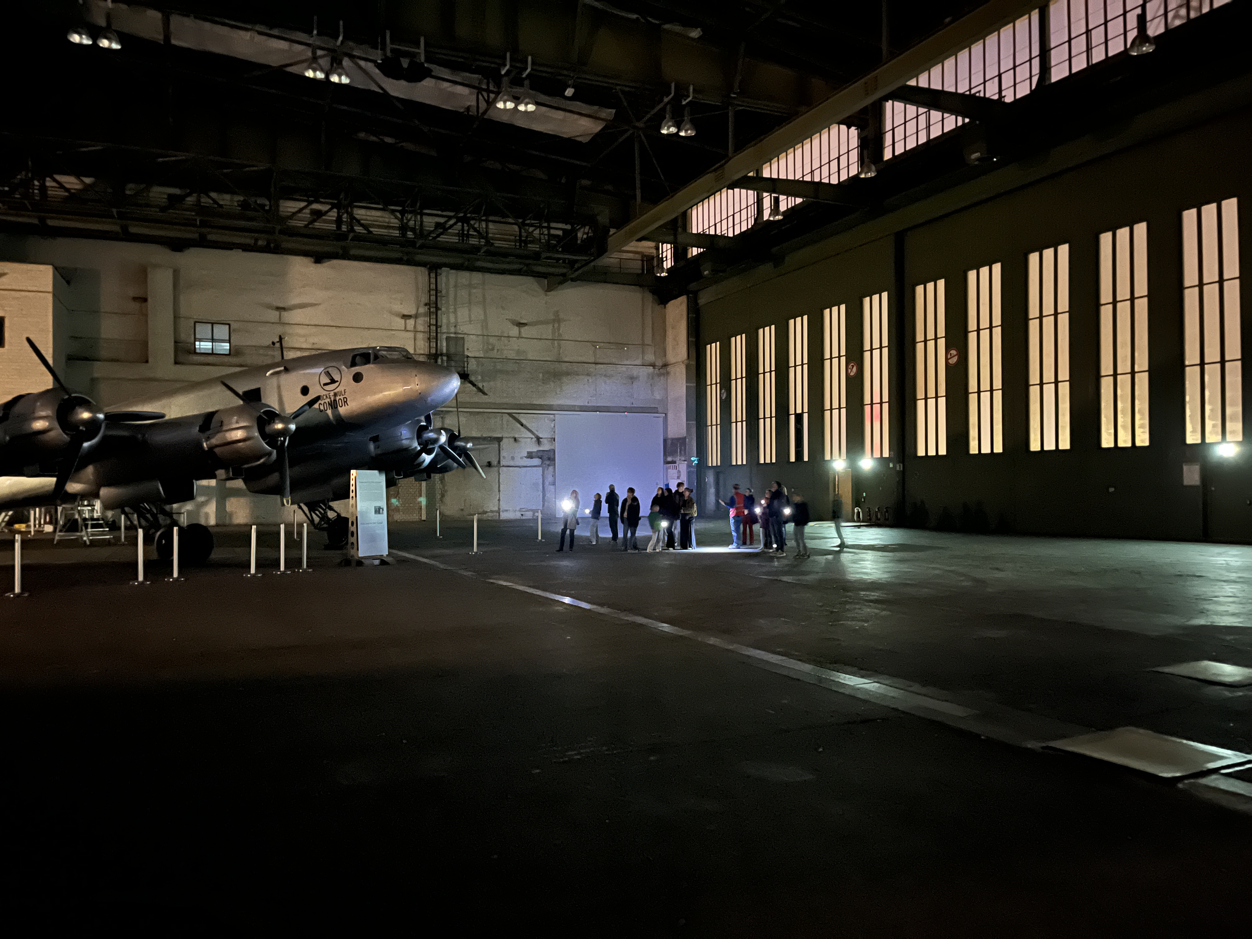 Kinder und Erwachsene leuchten bei einer Führung durch den Flughafen Tempelhof mit Taschenlampen auf ein Flugzeug, das in einem Hangar steht. 